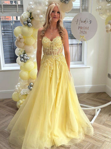A woman wearing a pale yellow strapless tulle prom dress with a lace-embellished bodice, standing indoors in front of a window and balloon decorations. A round sign reads 'Hadi's PROM 2026'.