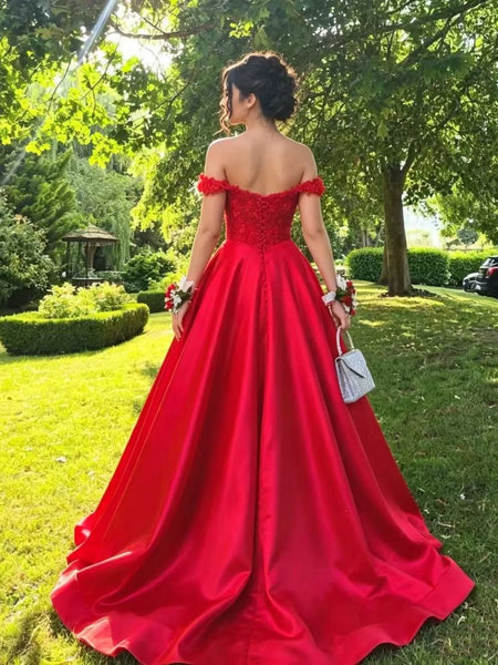 A woman stands outdoors in a lush green garden wearing a bright red off-the-shoulder ball gown with a fitted lace bodice and a full satin skirt. She is shown from the back, holding a small white handbag.